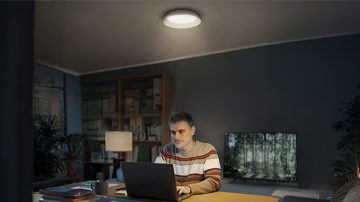 A man sits at a desk with a laptop, illuminated from above by a round ceiling light